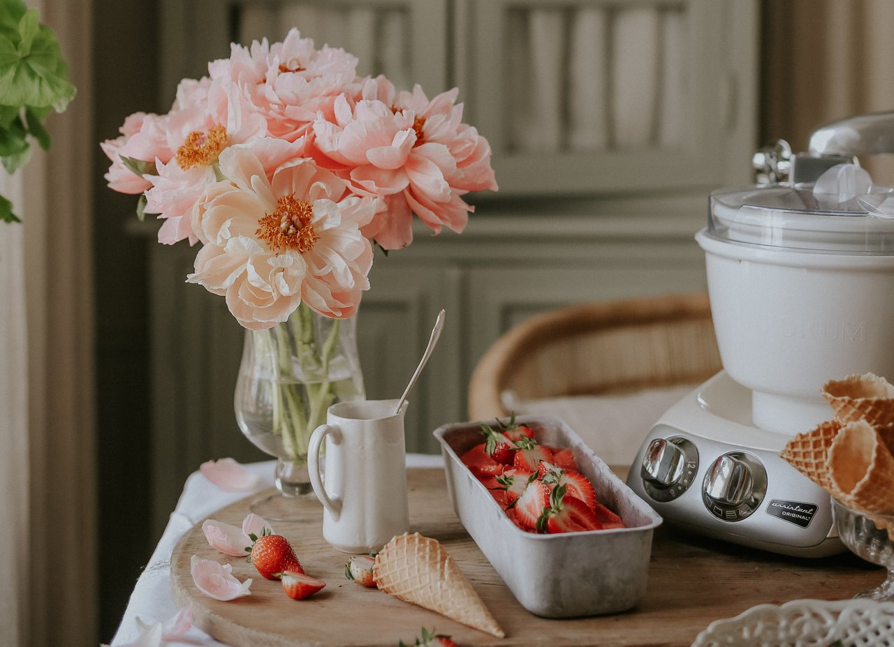 A bowl of strawberries and a vase of flowers on a table. On the table is an Ankarsrum Assistent Original in Cream, assembled with the Ice Cream Maker.