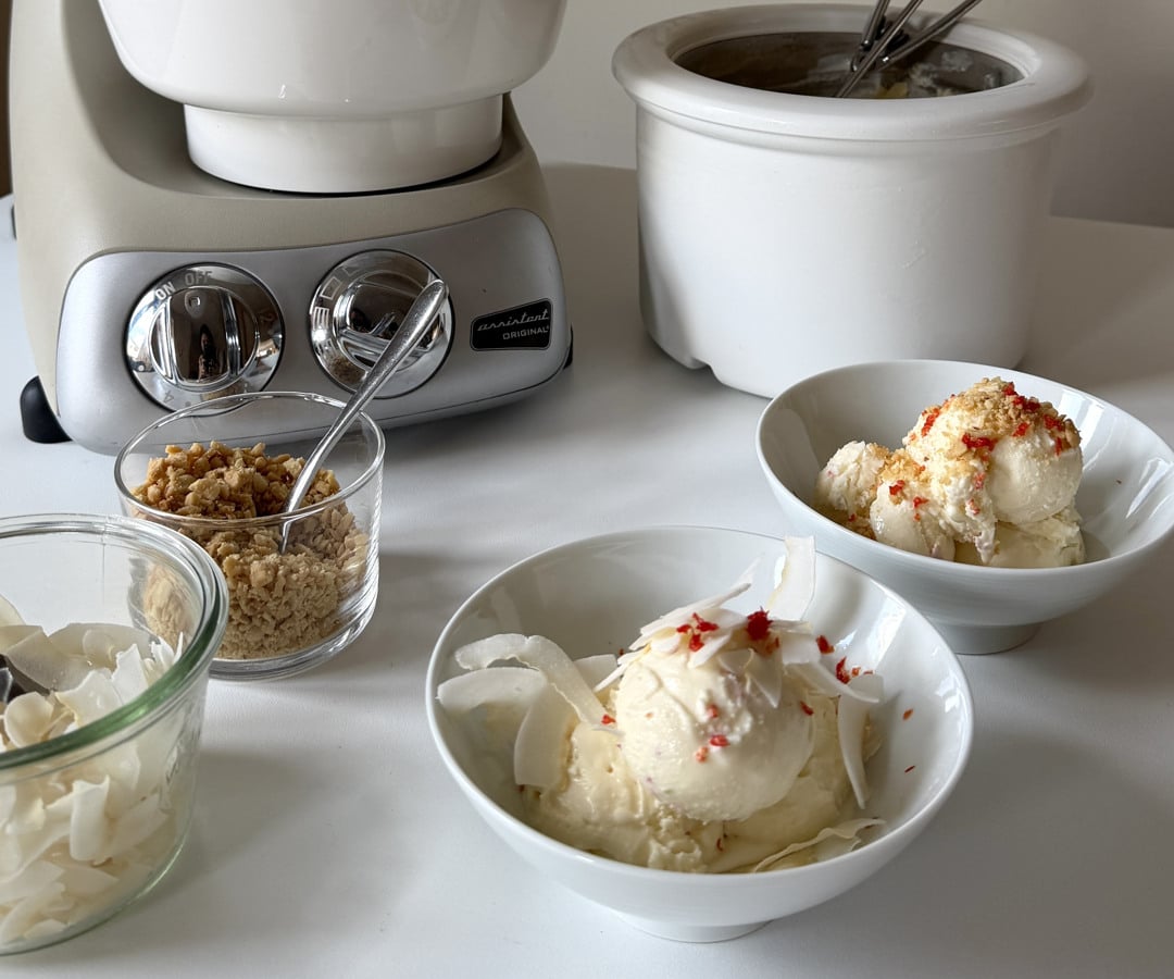 A white table topped with bowls of lime ice cream, garnished with chili and lemongrass. In the background, an Ankarsrum Assistent Original in Harmony Beige is fitted with the Ice Cream Maker attachment