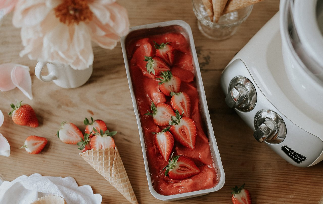 A bowl of strawberries and a vase of flowers on a table. On the table is an Ankarsrum Assistent Original in Cream, assembled with the Ice Cream Maker.