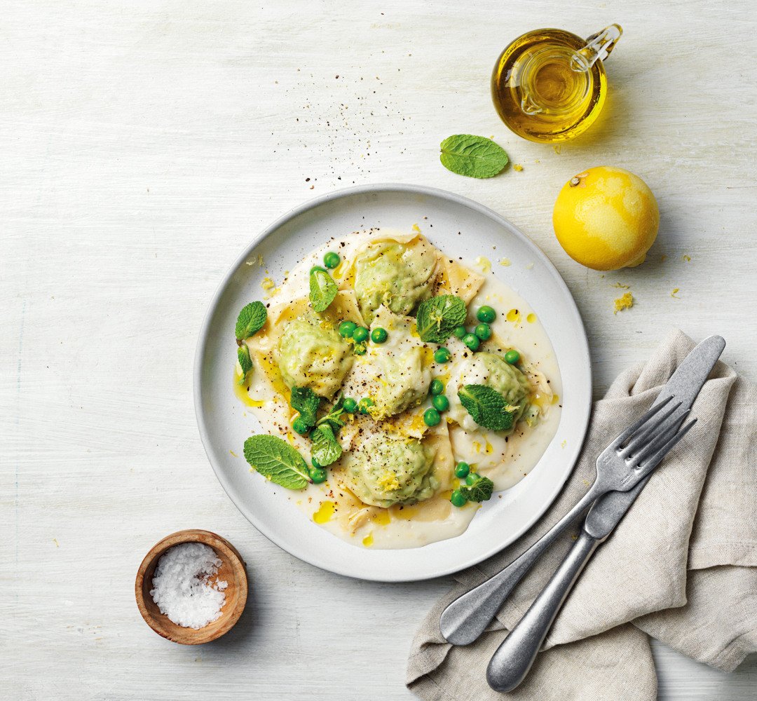 Ravioli with spinach and ricotta, topped with peas. Cutlery placed beside it, along with salt and a lemon.