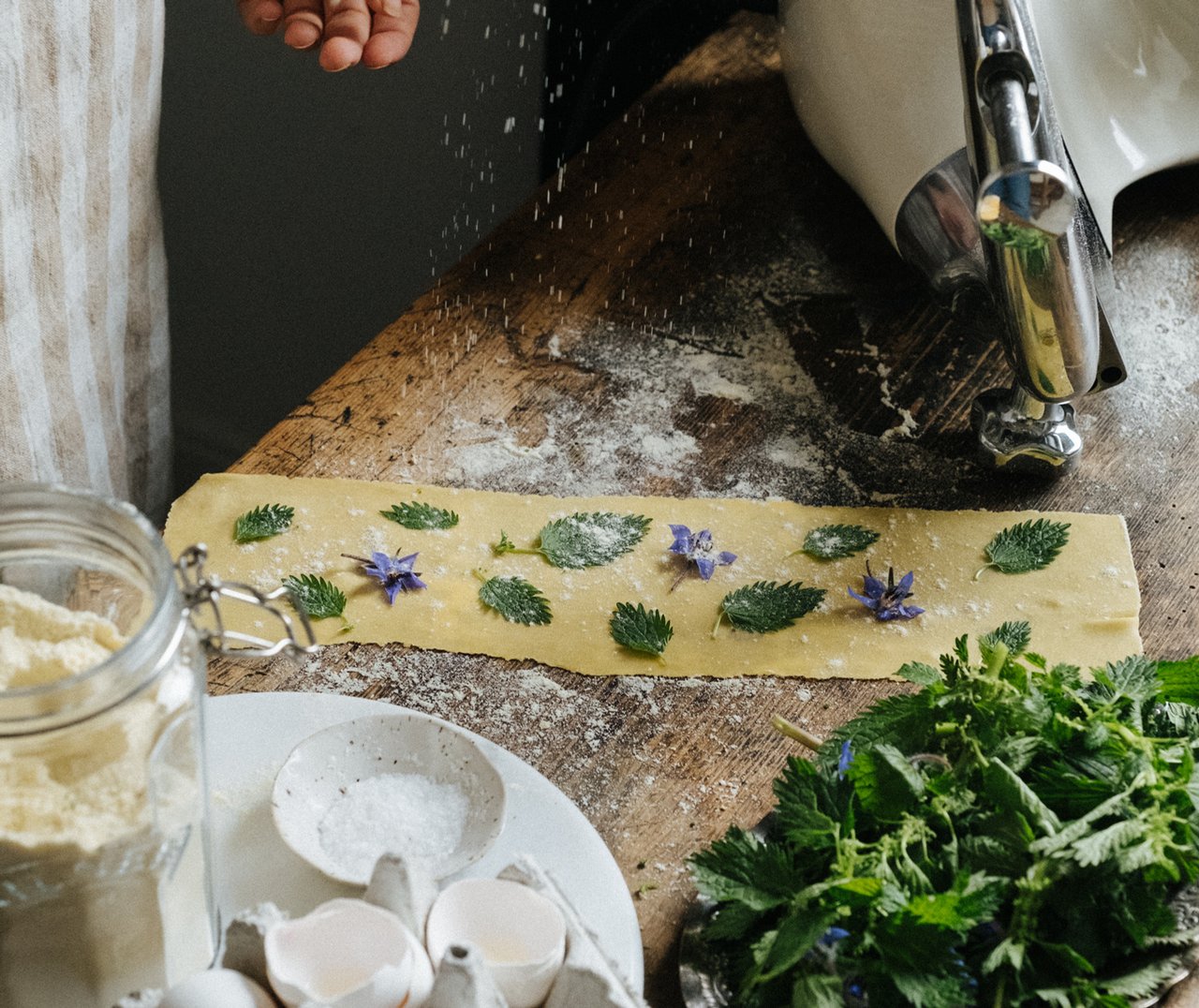 A person is making pasta at a dark wooden dining table, with flour scattered around the dough.