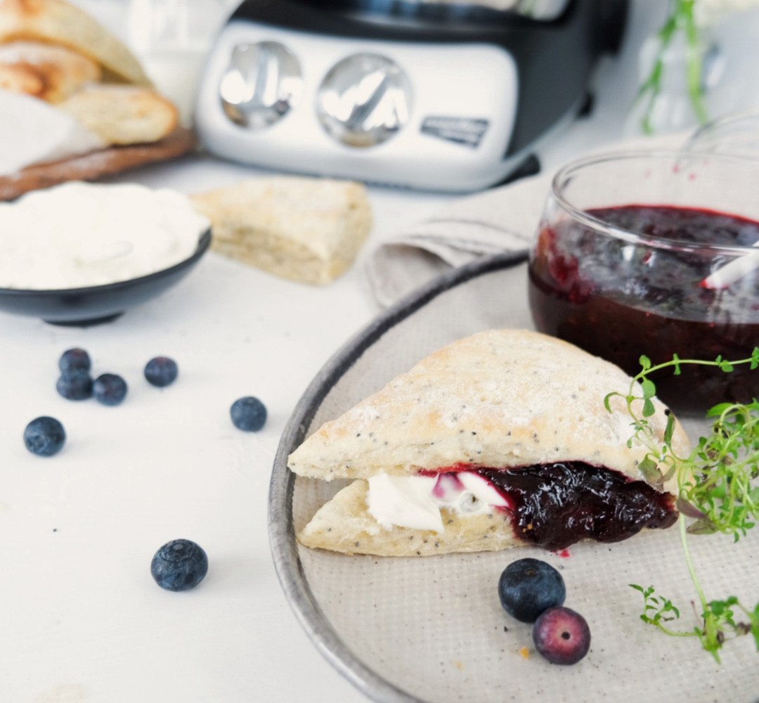 En tallrik med en scone fylld med sylt och en skål med sylt. En Ankarsrum Assistent i bakgrunden.