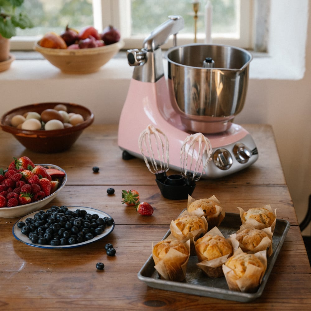 A woman is decorating cupcakes using a piping bag. On the table stands an Ankarsrum Assistent Original in Cloudy Pink, assembled with the Beater Bowl in Stainless Steel.