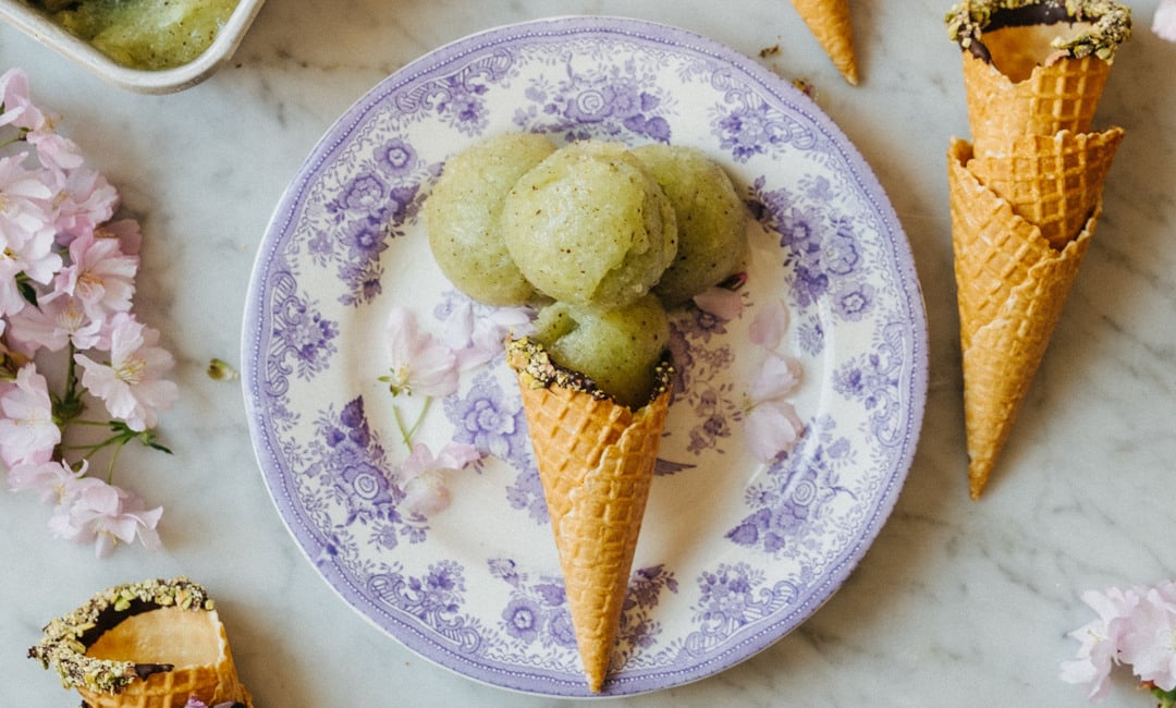 A person holding a plate with a kiwi sorbet cone. In the background, pink flowers are visible, along with a table holding a bowl of remaining ice cream.