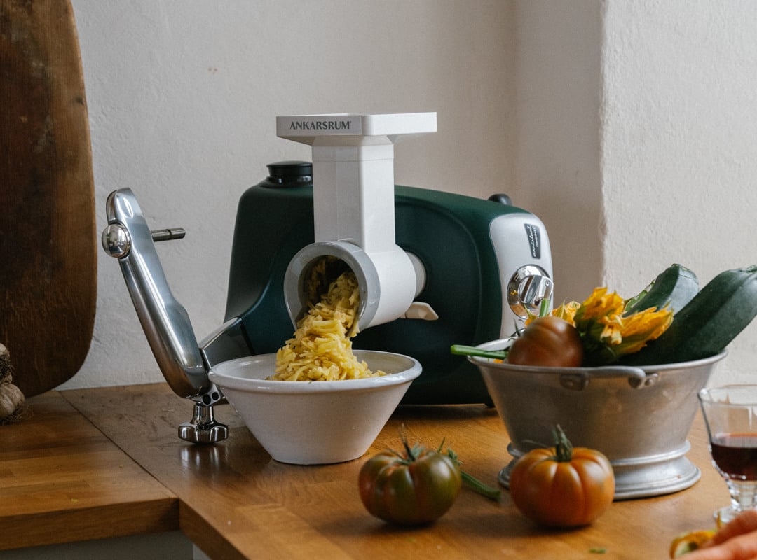 An Ankarsrum Assistent Original in Forest Green, fitted with the Vegetable Cutter attachment, stands on a wooden kitchen worktop surrounded by fresh vegetables and sliced courgette.