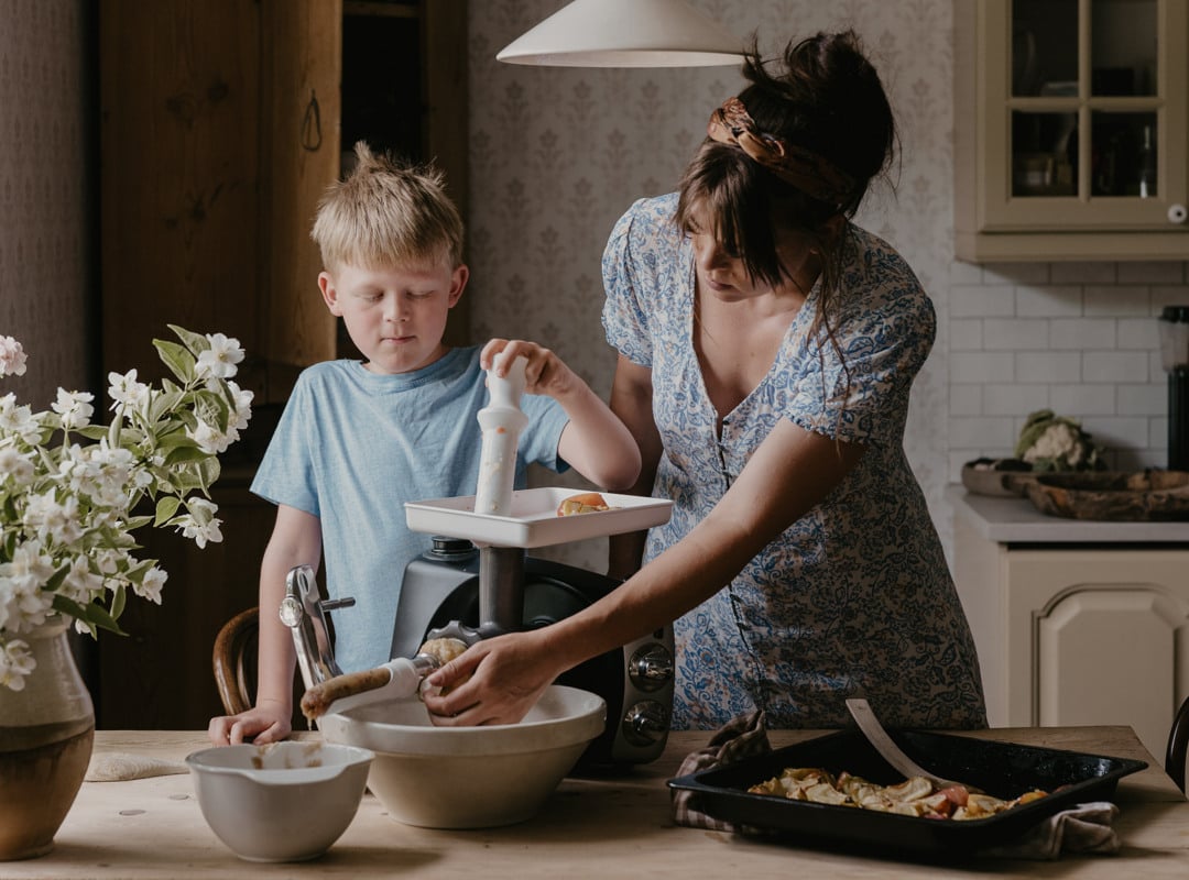 A woman and a boy use the Strainer attachment on an Ankarsrum Assistent Original in Forest Green to make fresh apple purée in a sunlit kitchen.