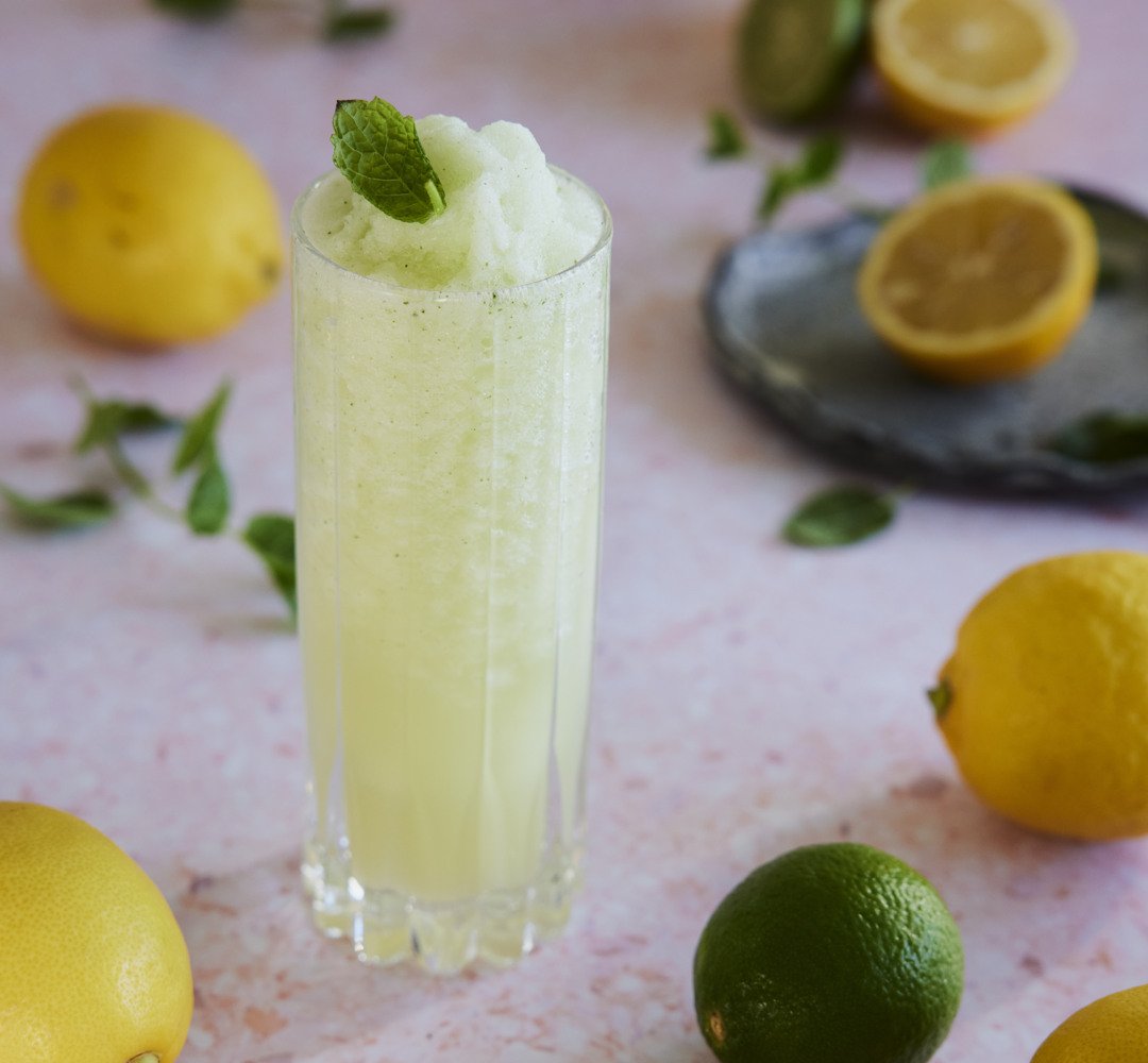 A glass of lemonade with a mint garnish and lemons around it.