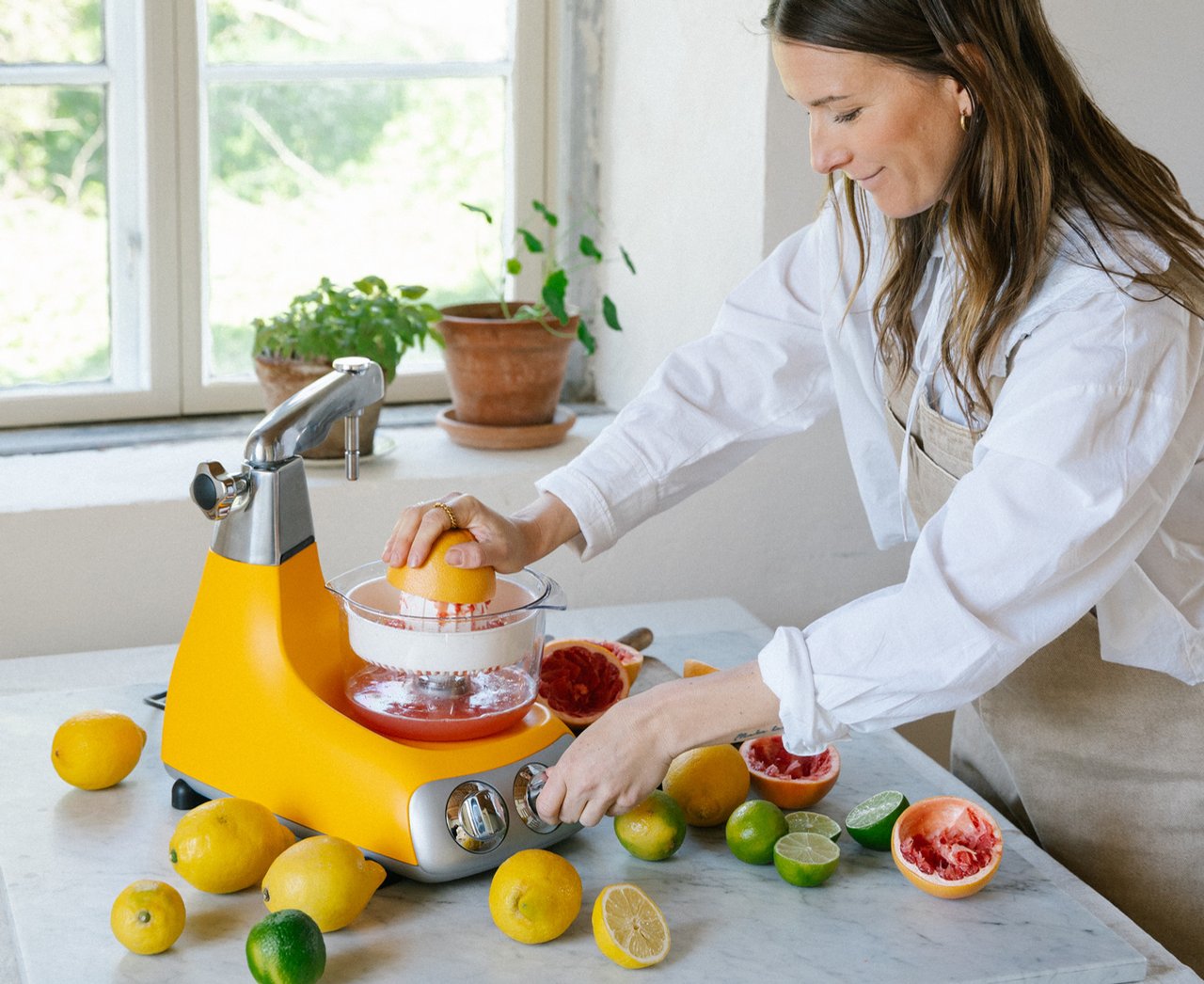 En kvinna använder en Ankarsrum Assistent Original med en citruspress för att pressa färsk grapefrukt i ett ljust kök. Citrons, limer och grapefrukt är ordnade på bänken, vilket framhäver förberedelsen av hemlagad juice.