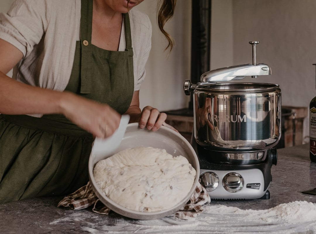 A woman prepares dough beside an Ankarsrum Assistent Original on a flour-dusted kitchen worktop.
