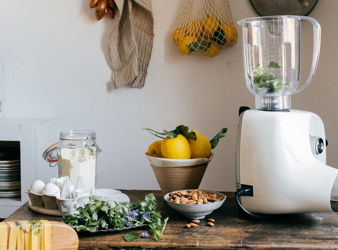 A blender is attached to a white Ankarsrum stand mixer sitting on top of a wooden table with vegetables in a bowl next to it.