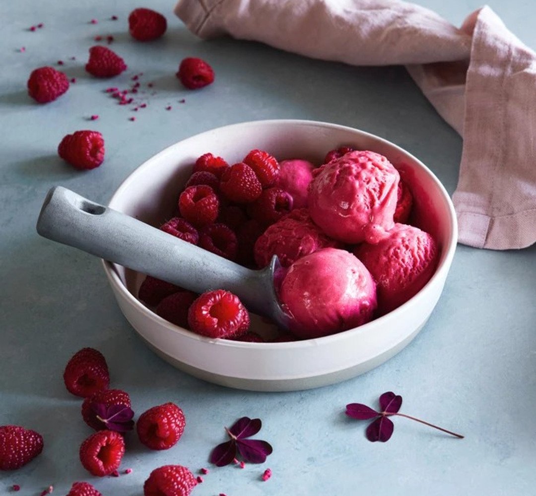 A bowl of raspberry ice cream with a spoon, with raspberries on the table beside it.