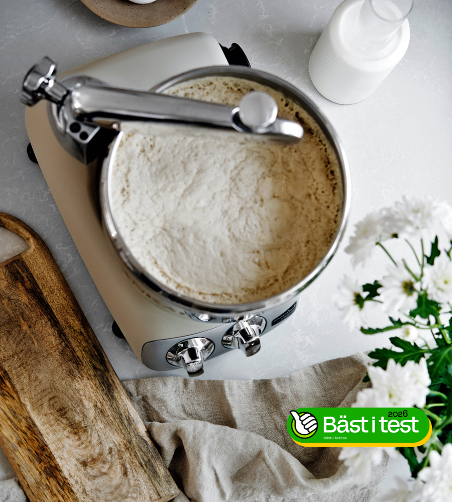 An Ankarsrum Assistent in Harmony Beige, kneading dough on a light-coloured kitchen worktop, with flowers, a cutting board and a kitchen towel beside it. Text reading “Best in Test” appears on one side.