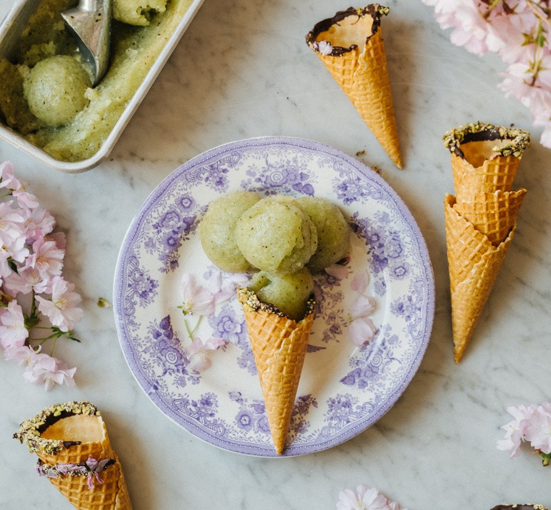 En person som håller en tallrik med en kiwi-sorbetkon. I bakgrunden syns rosa blommor, tillsammans med ett bord som har en skål med kvarvarande glass.