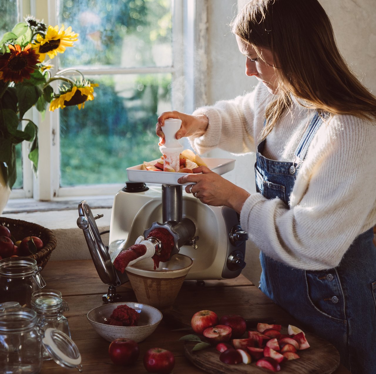 A woman uses the Strainer attachment on an Ankarsrum Assistent Original in Harmony Beige to make fresh apple purée in a sunlit kitchen.
