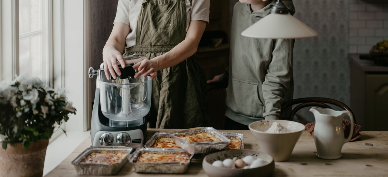 A person and a child are standing together by an Ankarsrum Assistent Original stand mixer in a bright kitchen. On the table in front of them are several freshly baked gratins, a bowl of flour, eggs, and a pitcher, creating a warm and cozy atmosphere of cooking together.