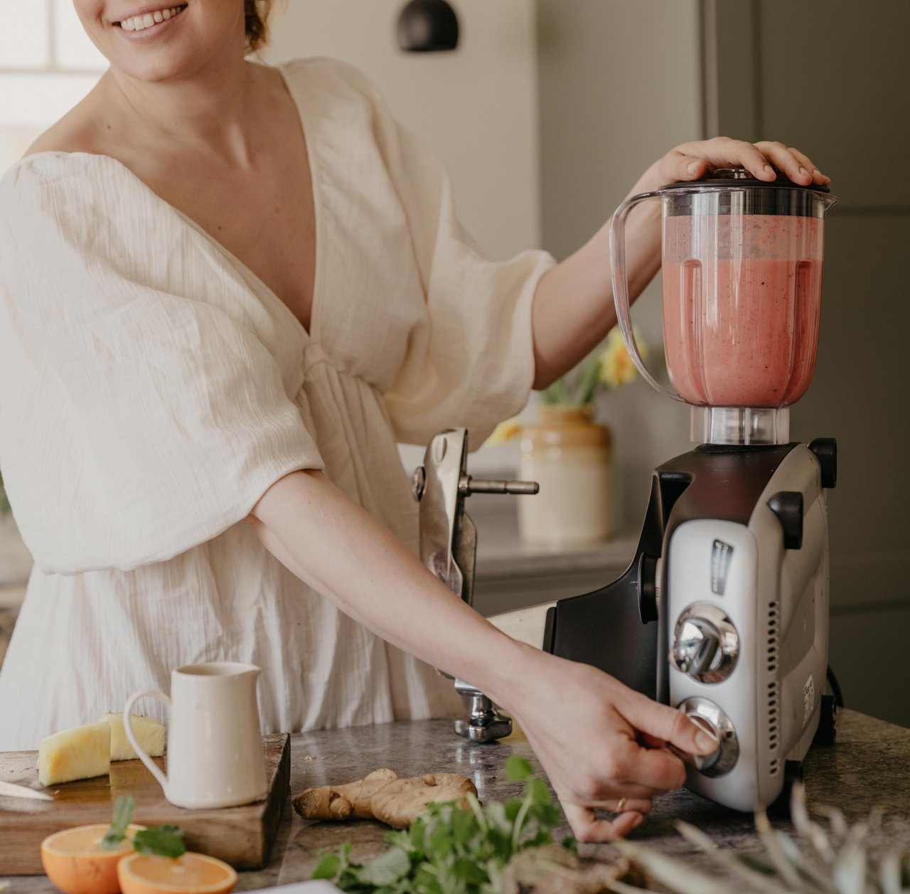 En person klädd i en vit blus använder Ankarsrum Assistent Original, med en mixerkanne fylld med rosa smoothie, och justerar maskinens hastighet. På köksbänken finns färsk ingefära, citrusfrukter, örter och en liten kanna som skapar en ljus och hemtrevlig atmosfär.