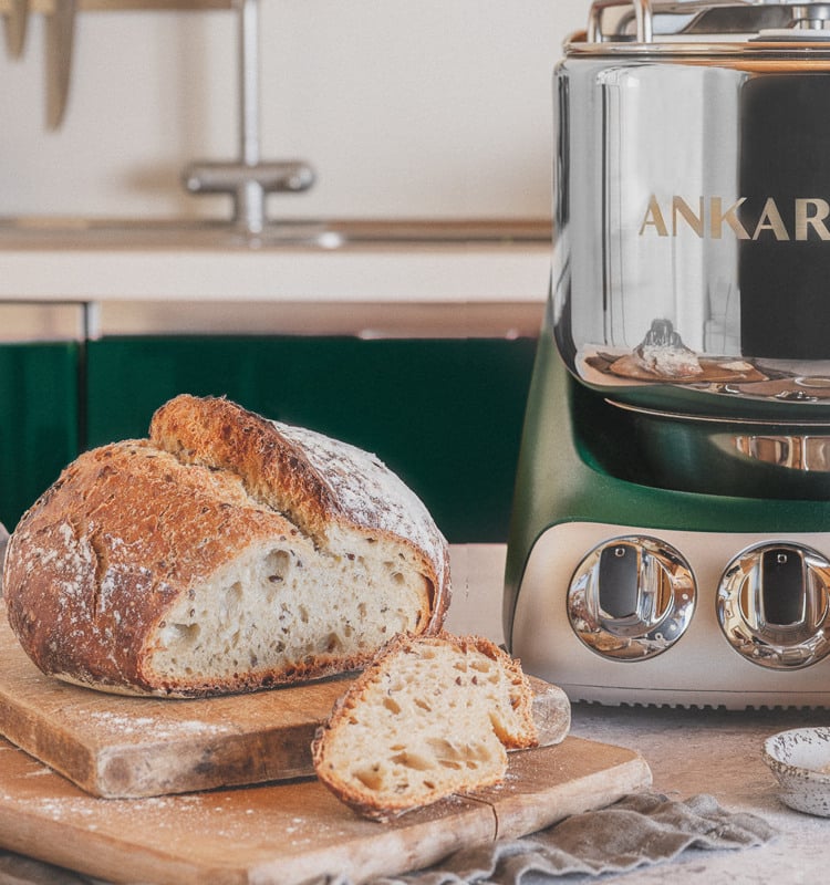 A loaf of bread sitting on top of a wooden cutting board. With an ankarsrum assistent on the side