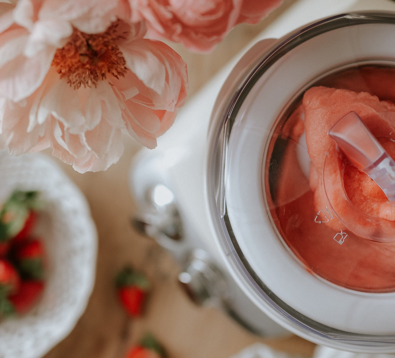 A bowl of strawberries and a vase of flowers on a table. On the table is an Ankarsrum Assistent Original in Cream, assembled with the Ice Cream Maker.
