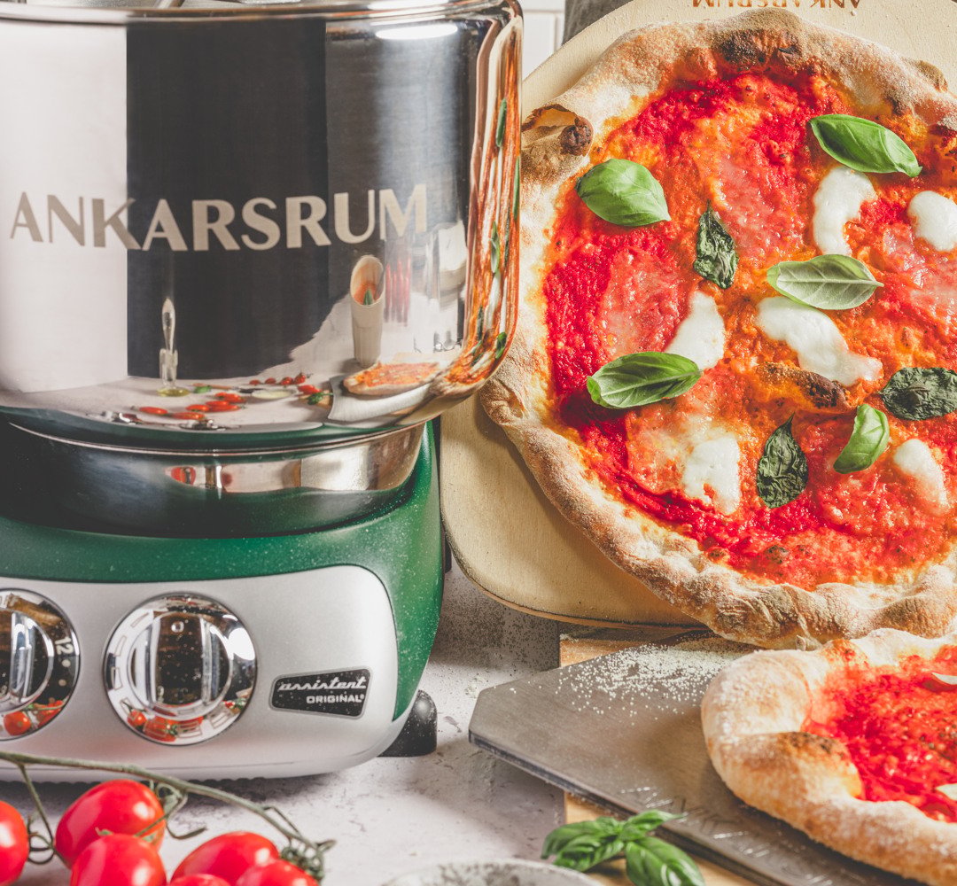 A pizza sitting on top of a pan on top of a counter. Ankarsrum assistent on the side
