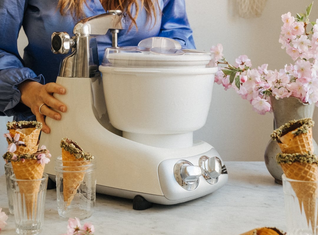 A woman is making ice cream with an Ankarsrum Assistent. Cones topped with melted chocolate are placed on the side.