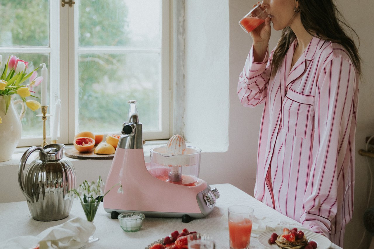 En kvinna klädd i en rosa pyjamas dricker ett glas färskpressad juice. Framför henne står ett frukostbord dukat med frukost, blommor och en Ankarsrum Assistent Original i färgen Cloudy Pink.