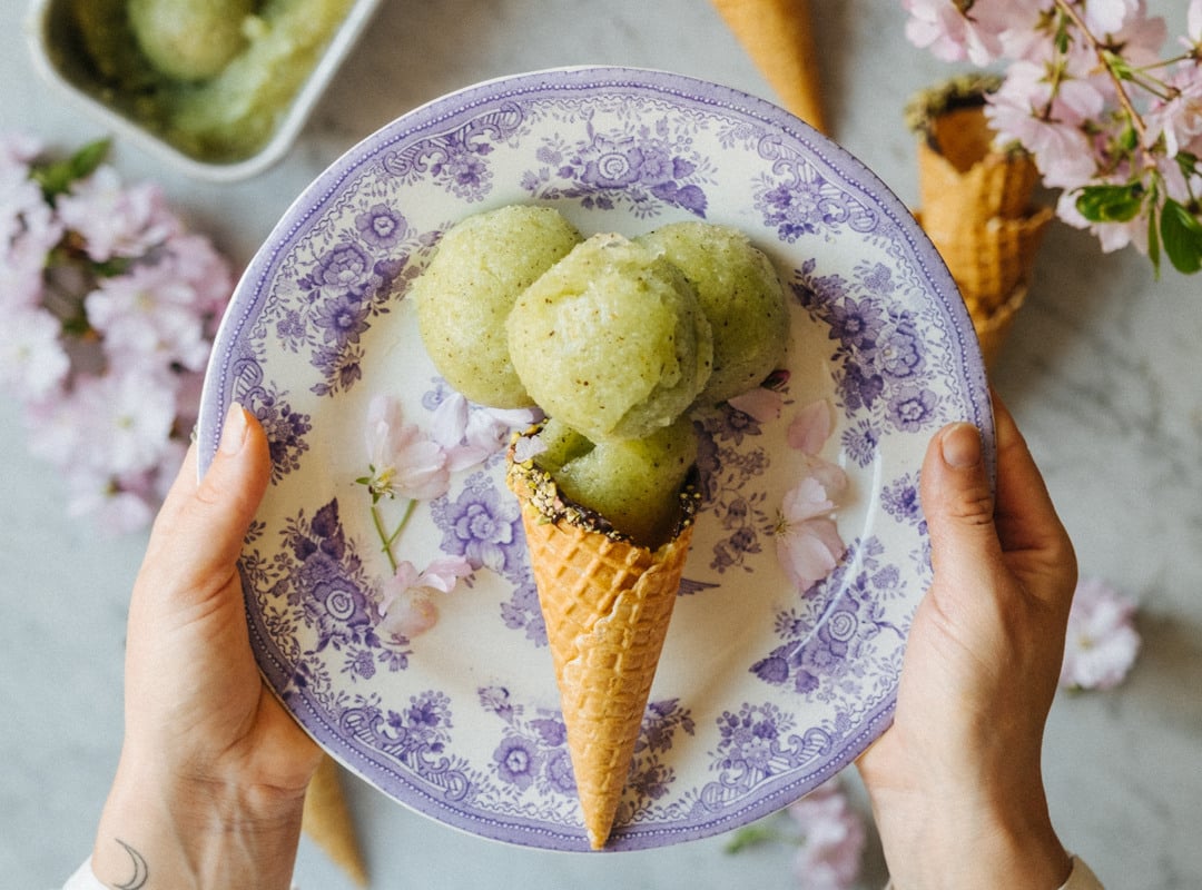 A person holding a plate with a kiwi sorbet cone. In the background, pink flowers are visible, along with a table holding a bowl of remaining ice cream.