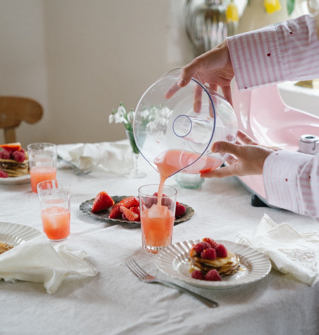 En person häller rosa juice från en Citruspress i ett glas fyllt med is. I bakgrunden syns ett dukat frukostbord med små pannkakor dekorerade med hallon, en tallrik med färska jordgubbar, glas med juice, vita tygservetter och en liten vas med vita blommor. Den ljusa miljön skapar en varm och inbjudande atmosfär.