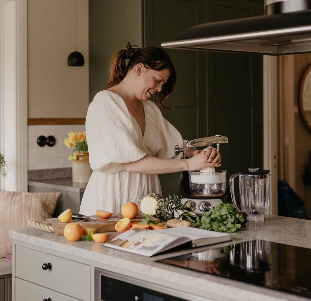 En kvinna klädd i vit klänning använder Ankarsrum Assistent Original med Citruspressen för att pressa färsk citrusjuice. På köksbänken ligger apelsiner, citron, ananas, ingefära, färska örter och en öppen kokbok, och bredvid köksmaskinen står en tom mixerkanna.