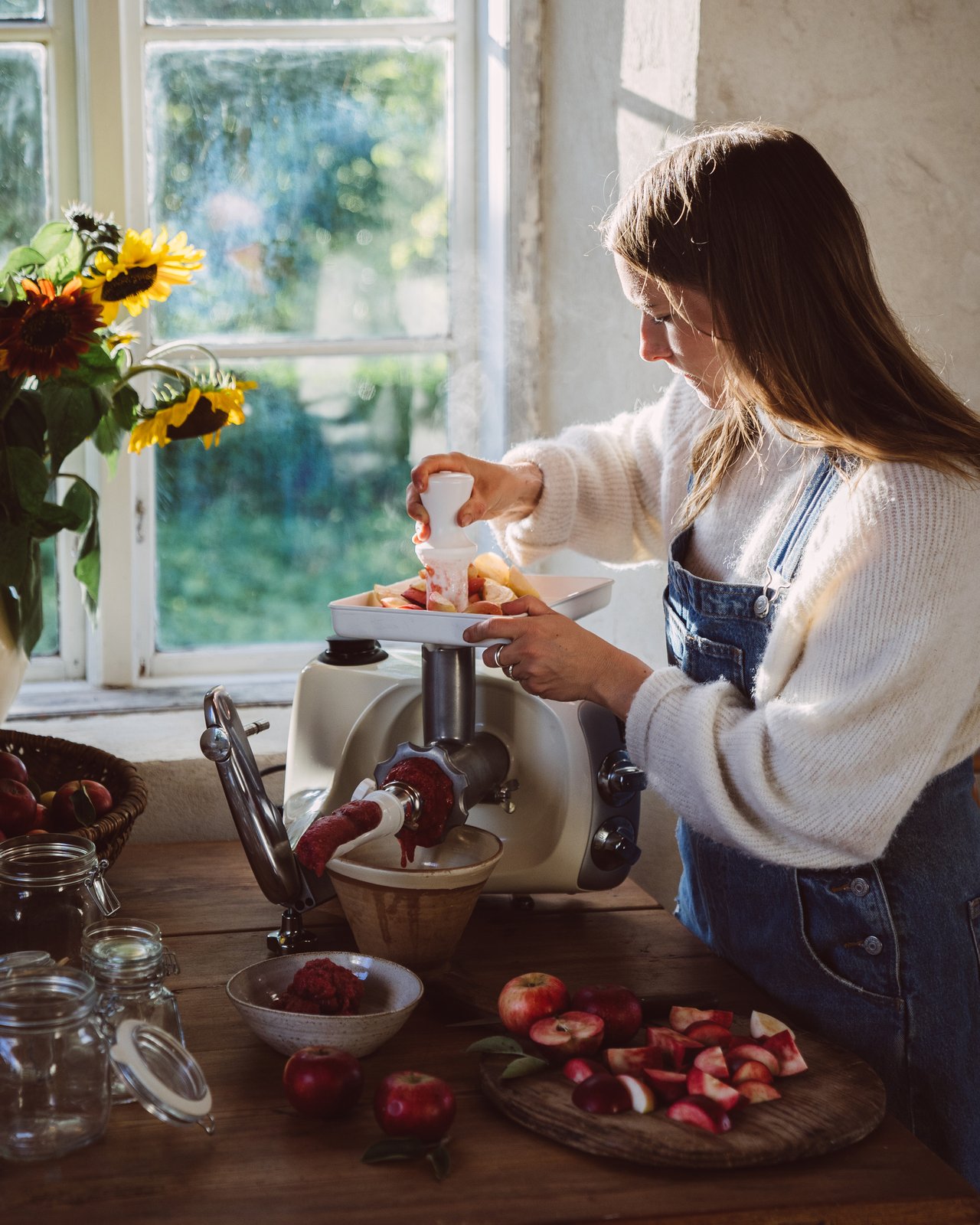 A woman uses the Strainer attachment on an Ankarsrum Assistent Original in Harmony Beige to make fresh apple purée in a sunlit kitchen.
