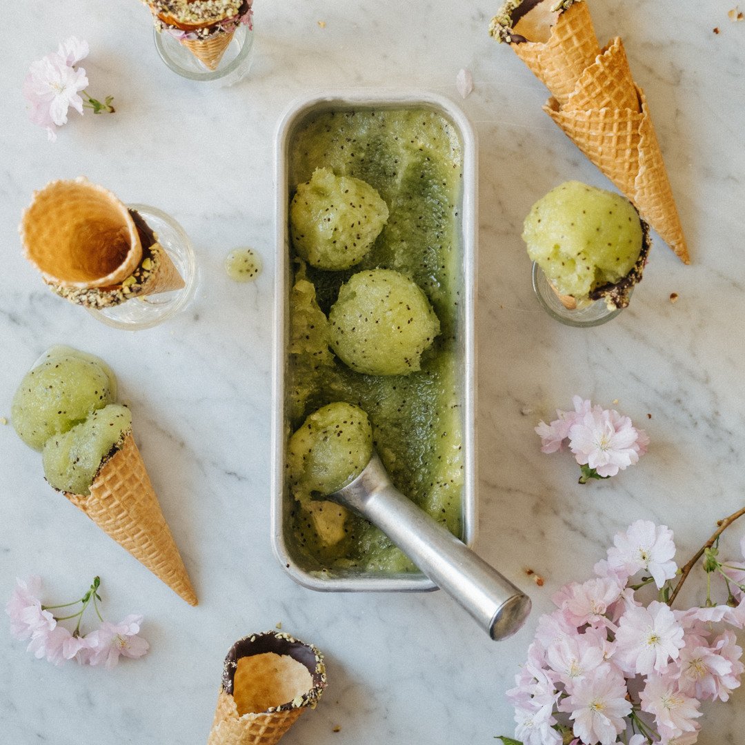 Kiwi-sorbet i en metallform, omgiven av glasskoner, pistagenötter, maränger och rosa blommor på en ljus marmor yta.