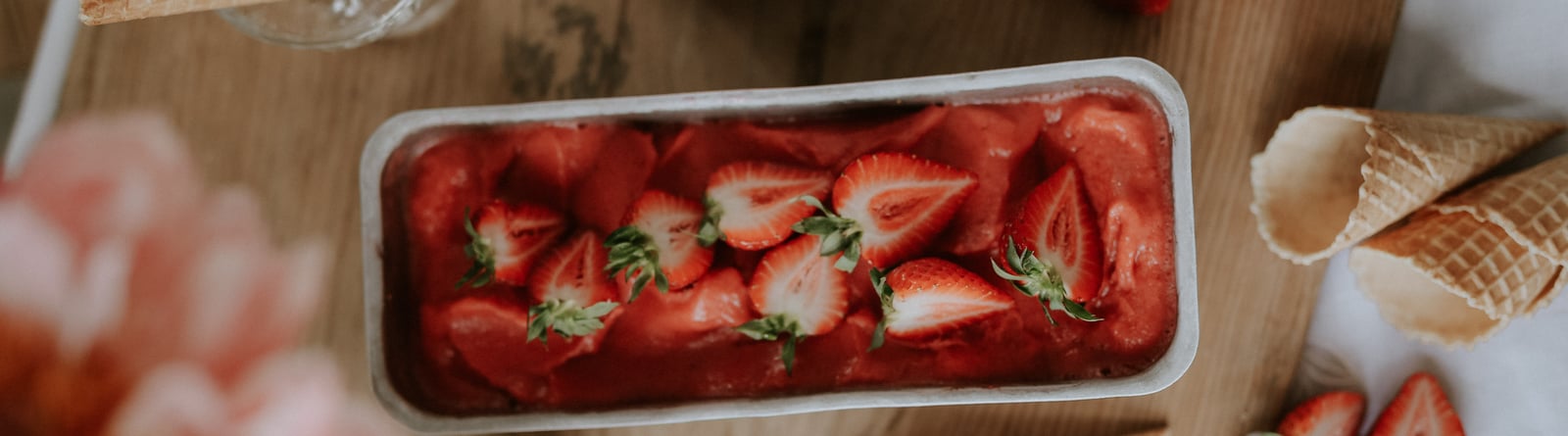 Strawberry sorbet in a bowl on a wooden table, surrounded by strawberries and ice cream cones.