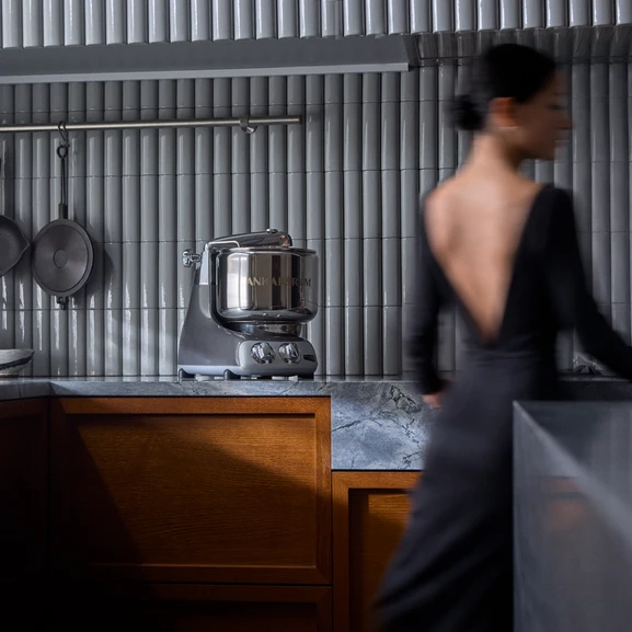 Ankarsrum Assistent Original nel colore Black Chrome su un piano di lavoro, vista da lontano, con una donna in movimento accanto alla macchina in cucina.