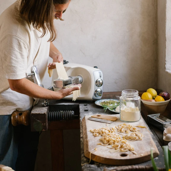 En person laver fettuccine på en køkkenbordplade i træ, med et meldrysset skærebræt og frisklavet fettuccine ved siden af.
