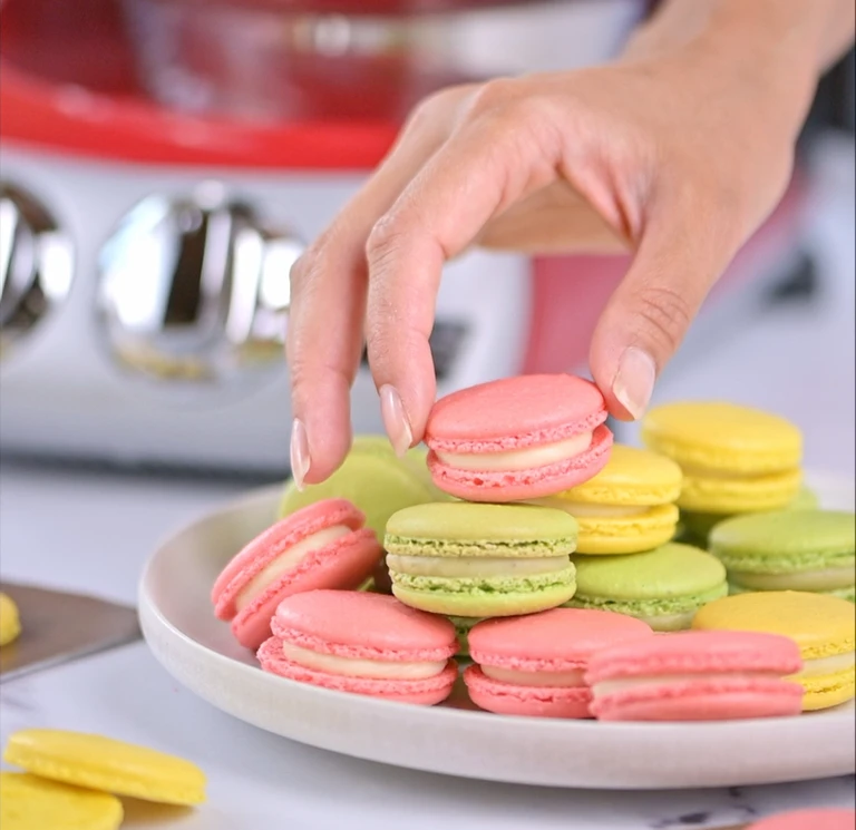 Hands holding pink and green macarons in a kitchen setting.