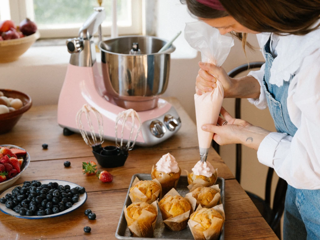 Beste kjøkkenmaskinen i fargen Cloudy Pink brukes til å sprøyte frosting på muffins på kjøkkenbord