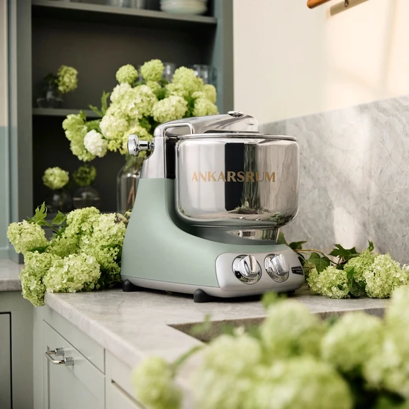 Ankarsrum Assistent Original in the color Nordic Sage on a marble countertop in a bright kitchen, surrounded by white hydrangea flowers.