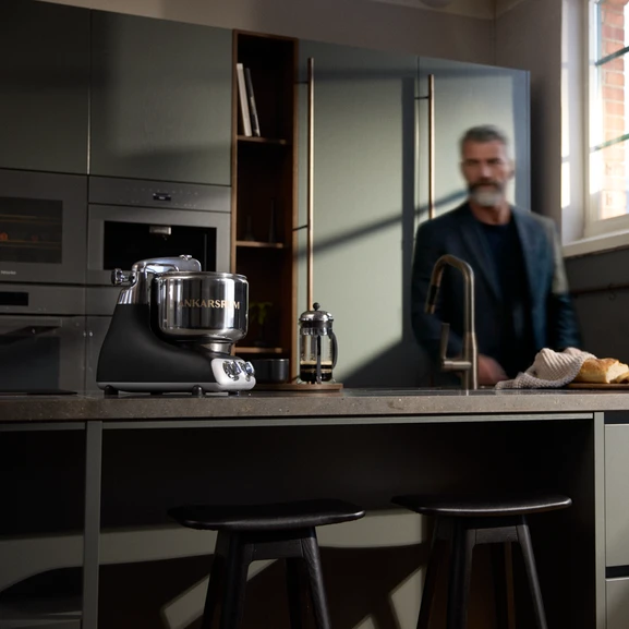 Ankarsrum Assistent Original in Black on a kitchen island in a dark kitchen, with a man visible in the background.