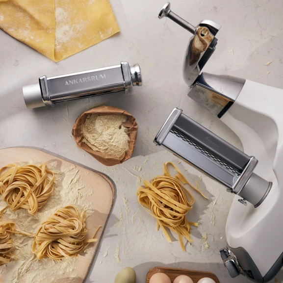 Ankarsrum Assistent Original mit montierten Pastaschneidern, mit Mehl und Nestern aus frisch zubereiteter Pasta daneben.