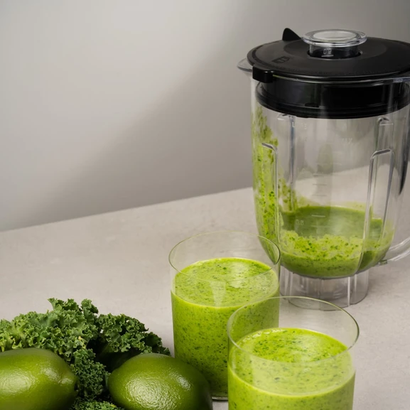 Blender på en kjøkkenbenk med to glass grønn smoothie ved siden av.