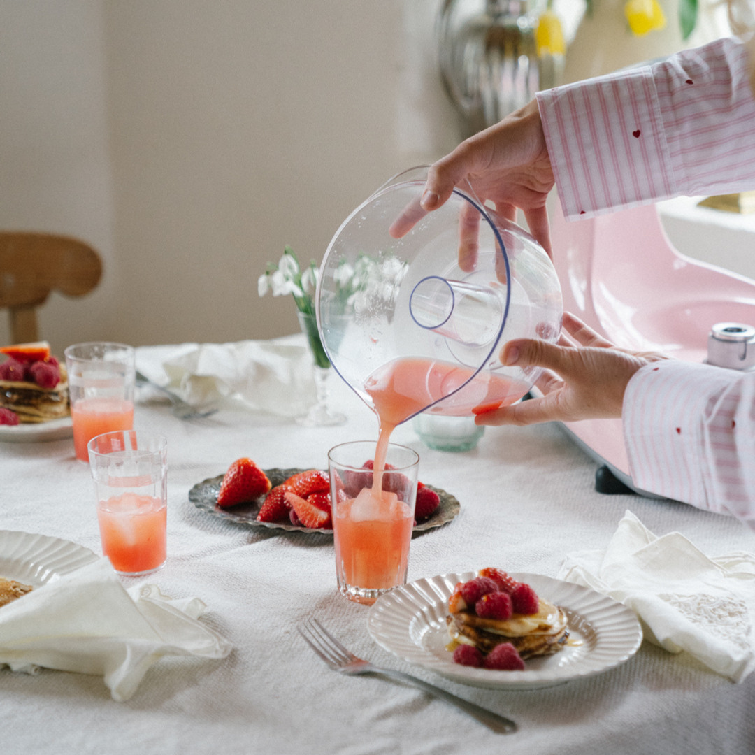 En person hælder en lyserød jordbærdrik op i et glas ved et dækket bord med friske bær, pandekager og flere glas med drikkevarer. En Ankarsrum Assistent Original er synlig i baggrunden.