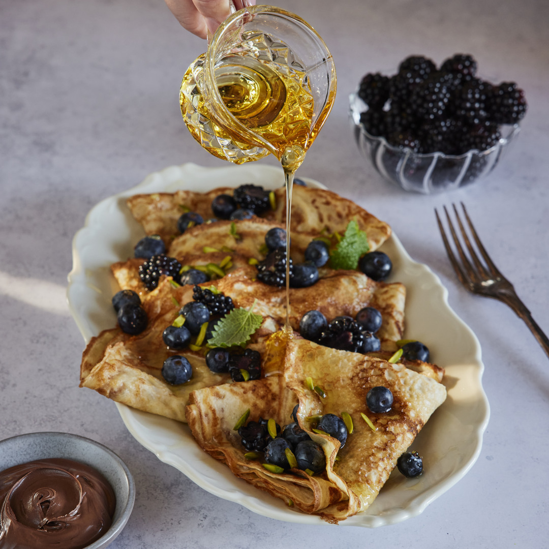 Honning hældes over foldede crêpes toppet med blåbær, brombær og pistacienødder på et serveringsfad. Ved siden af står en skål med brombær og en skål med chokoladecreme.