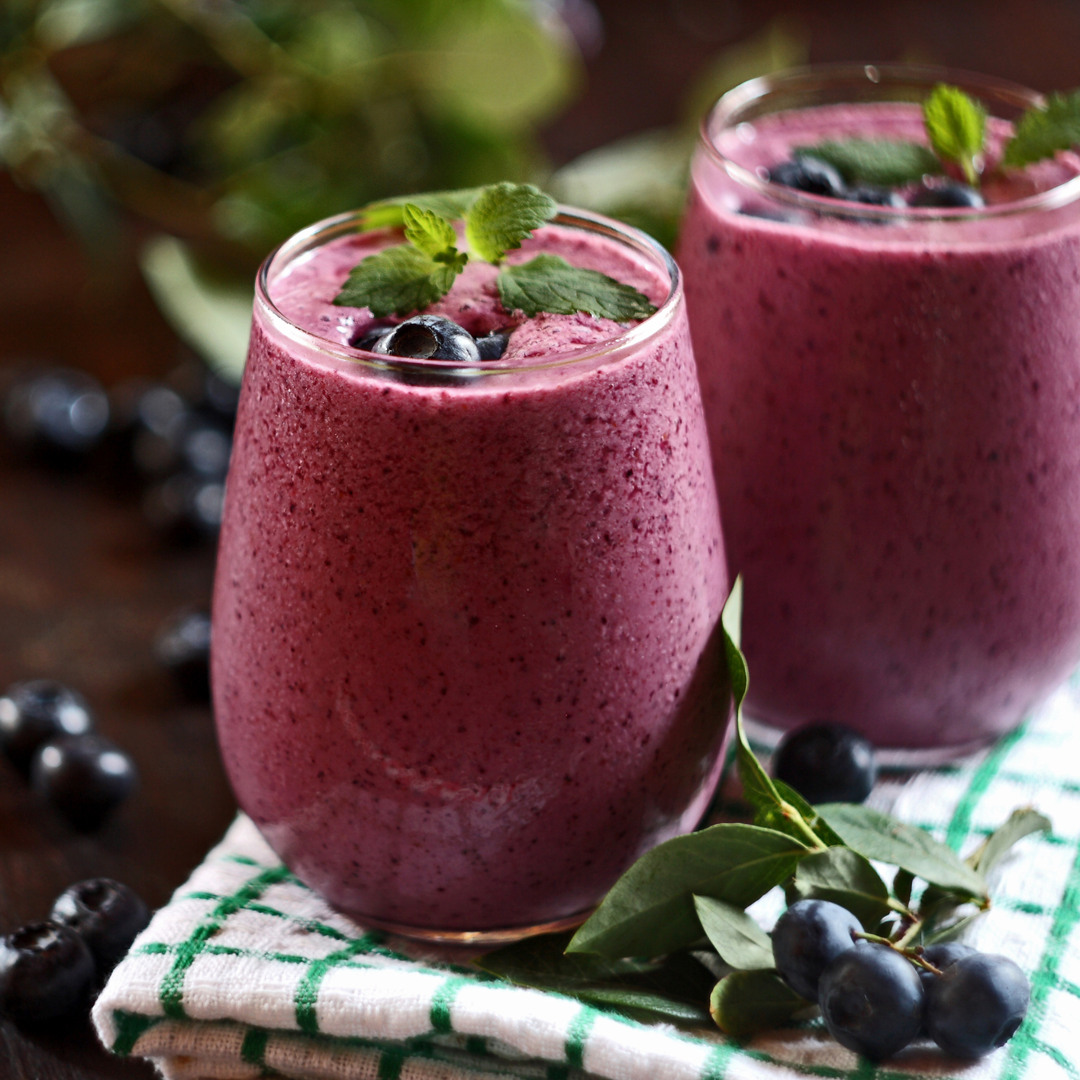 To glass med lilla blåbærsmoothie garnert med friske blåbær og mynteblader. Blåbær og grønne blader ligger rundt glassene på et bord med et kjøkkenhåndkle i forgrunnen.
