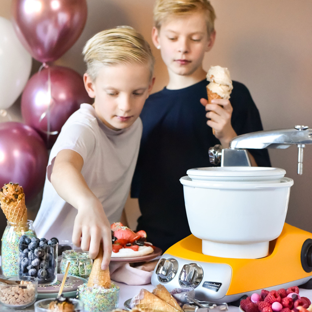 Två barn som gör glass med Ankarsrums assistent tillsammans med tillbehöret glassmaskin. De har glasstrutar, strössel och frukt vid sidan om att toppa deras glass med.