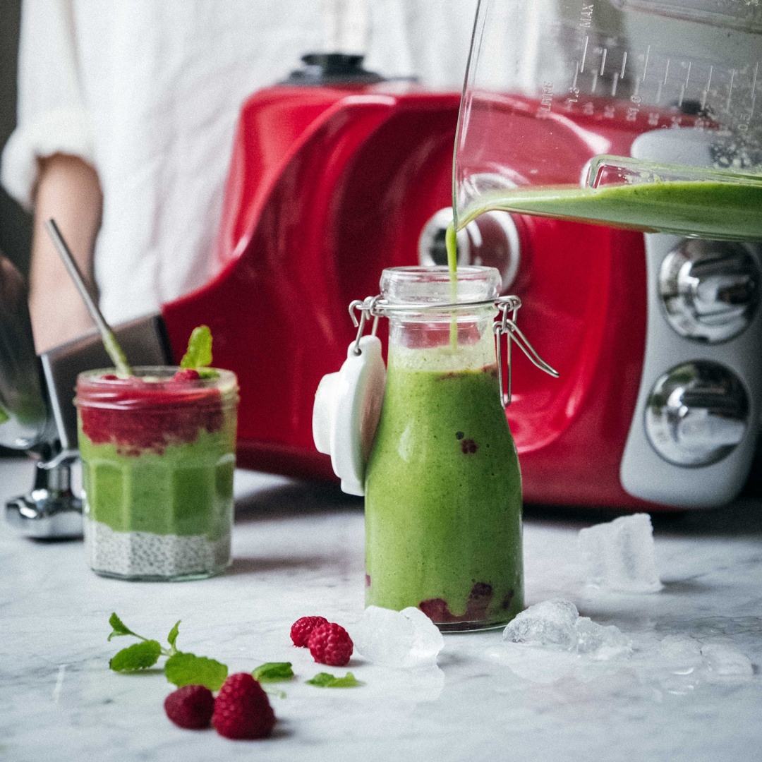 En grøn smoothie hældes i en glasflaske foran en Ankarsrum Assistent Original ved siden af en chiapudding toppet med hindbær.