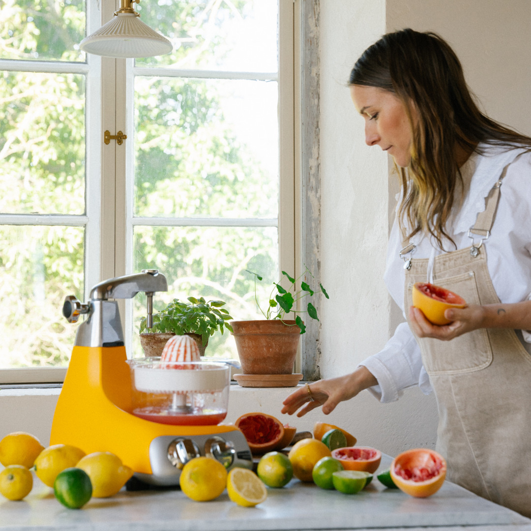 En kvinde bruger Ankarsrums citruspresser, monteret på en Ankarsrum Assistent Original i farven Sunbeam Yellow, til at presse frisk citrusjuice. Citroner, limefrugter og grapefrugter ligger på køkkenbordpladen.