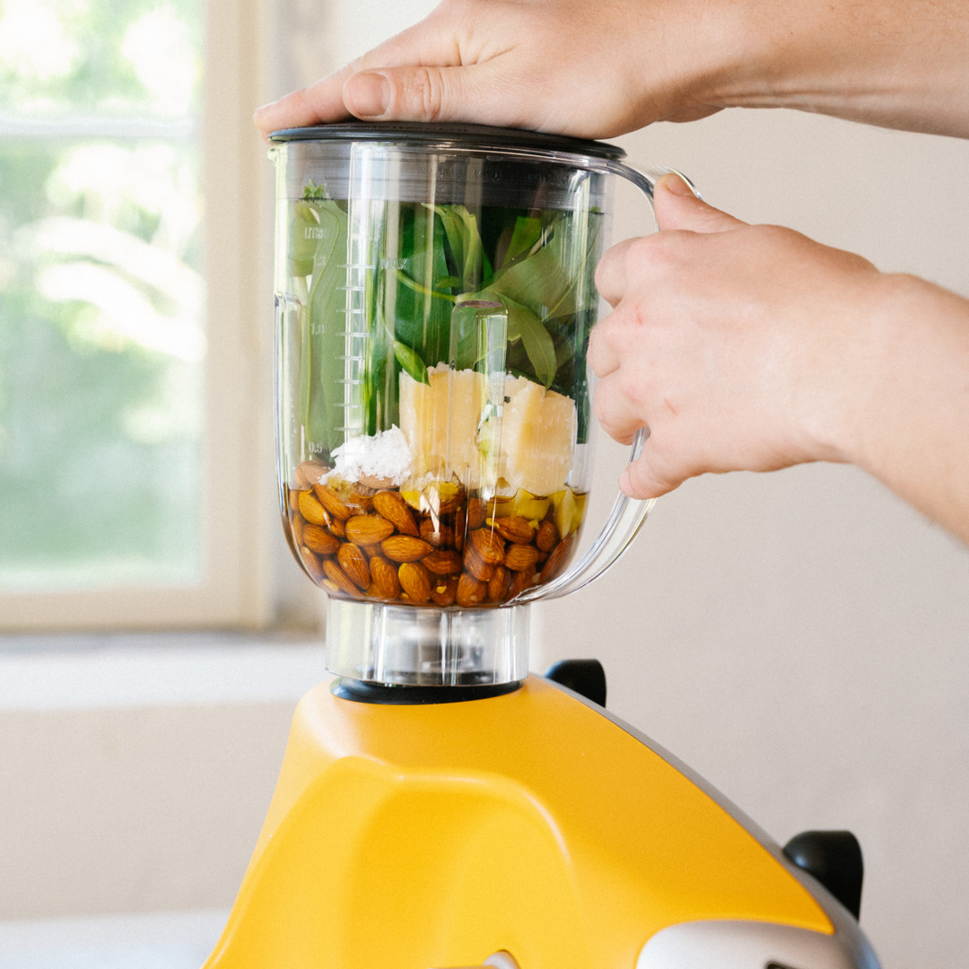 Nærbillede af Ankarsrums blender fyldt med friske ingredienser som spinat, mandler, parmesan og olivenolie, monteret på en Ankarsrum Assistent Original i farven Sunbeam Yellow.