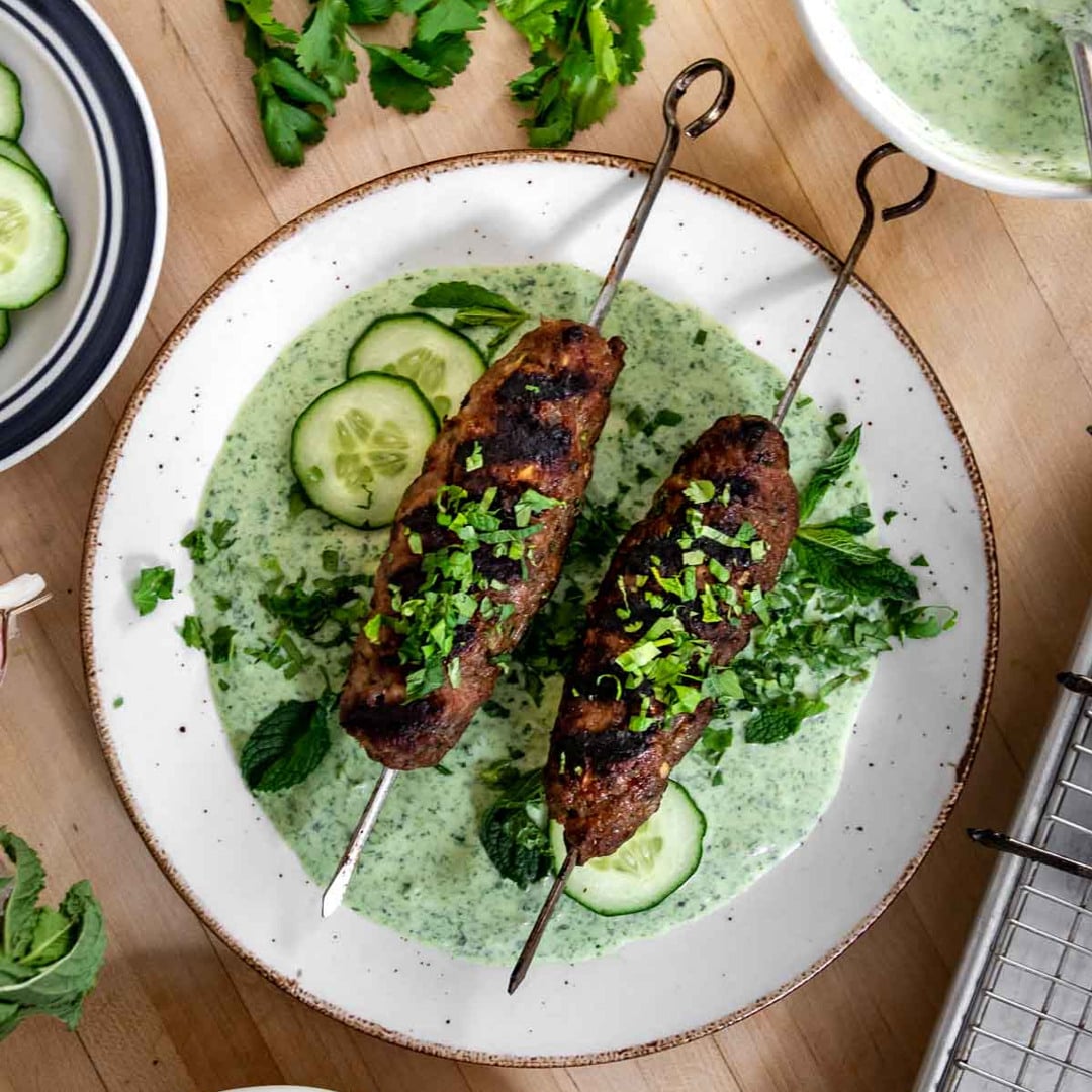 To grillede kebabspyd serveret med krydret yoghurtsauce, agurk og friske krydderurter på et hvidt fad.