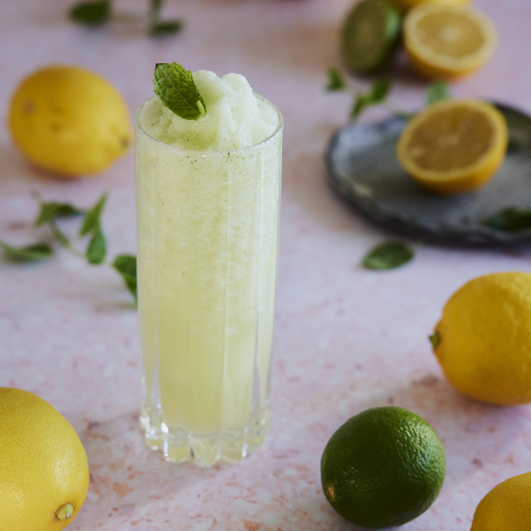 Tall glass filled with a pale yellow citrus slush topped with a fresh mint leaf. Lemons, limes, and mint are scattered around the glass on a light-colored surface.