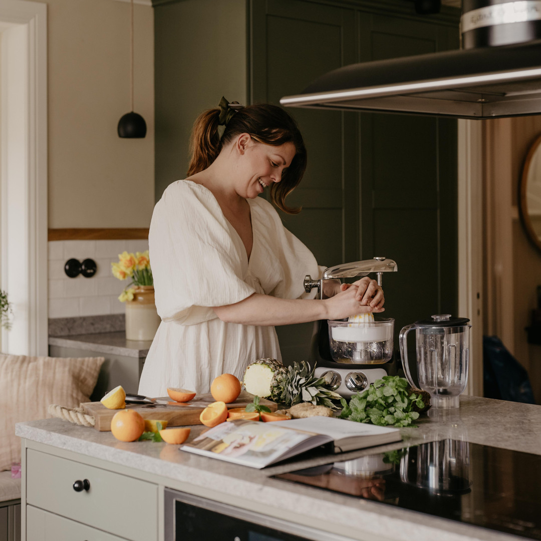 Person using the Ankarsrum Assistent Original with a citrus press attachment to juice citrus fruits on a kitchen island. Fresh oranges, pineapple, ginger, and herbs are displayed beside an open recipe book in a modern kitchen.